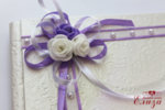 Сватбена книга за пожелания с твърди корици и украсена в лилаво със сатенени панделки и изкуствени цветчета.